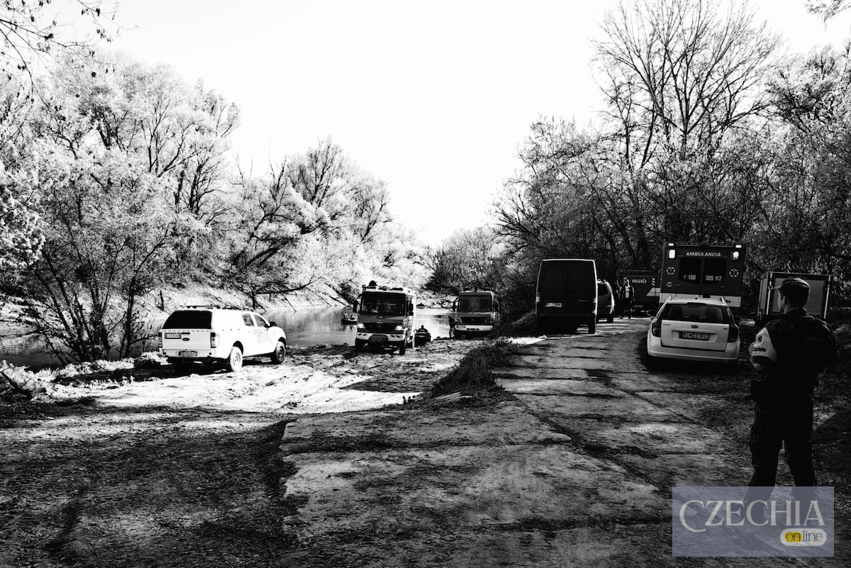 Чорно-біле фото: машини швидкої допомоги, пожежники та поліція на березі річки шукають людей.