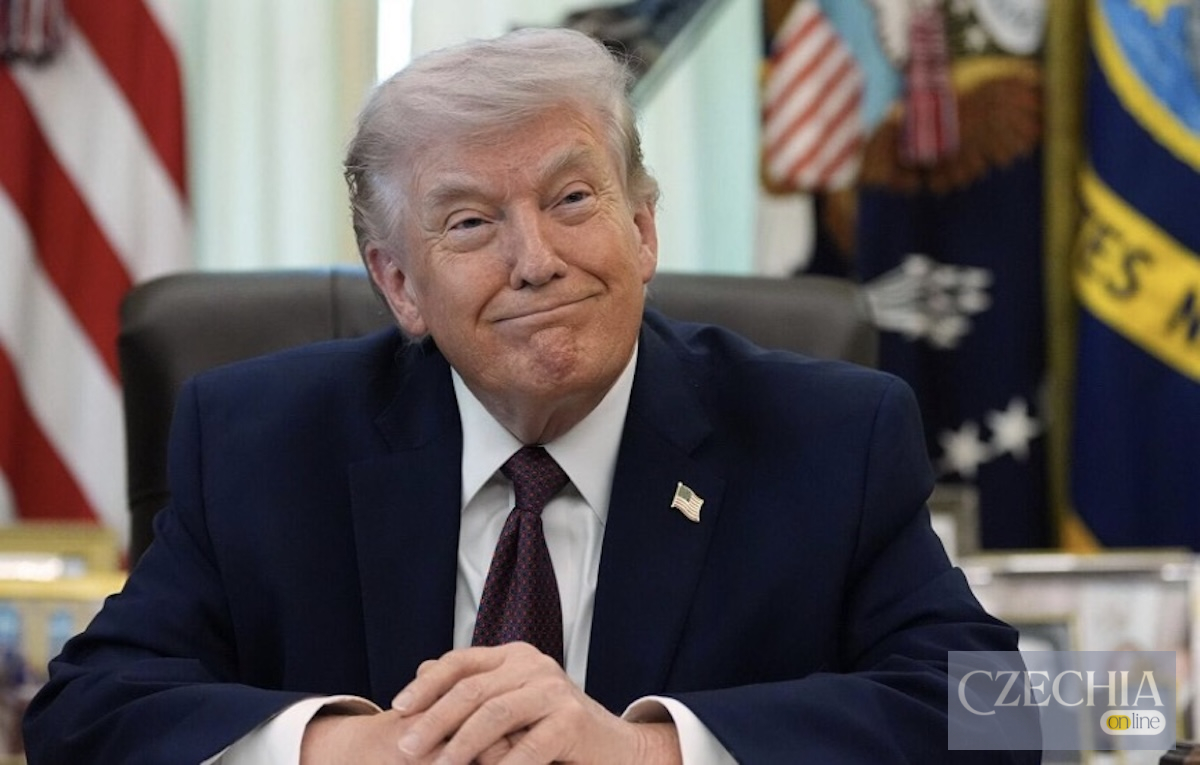 Усміхнений Дональд Трамп сидить за столом на тлі американських прапорів.