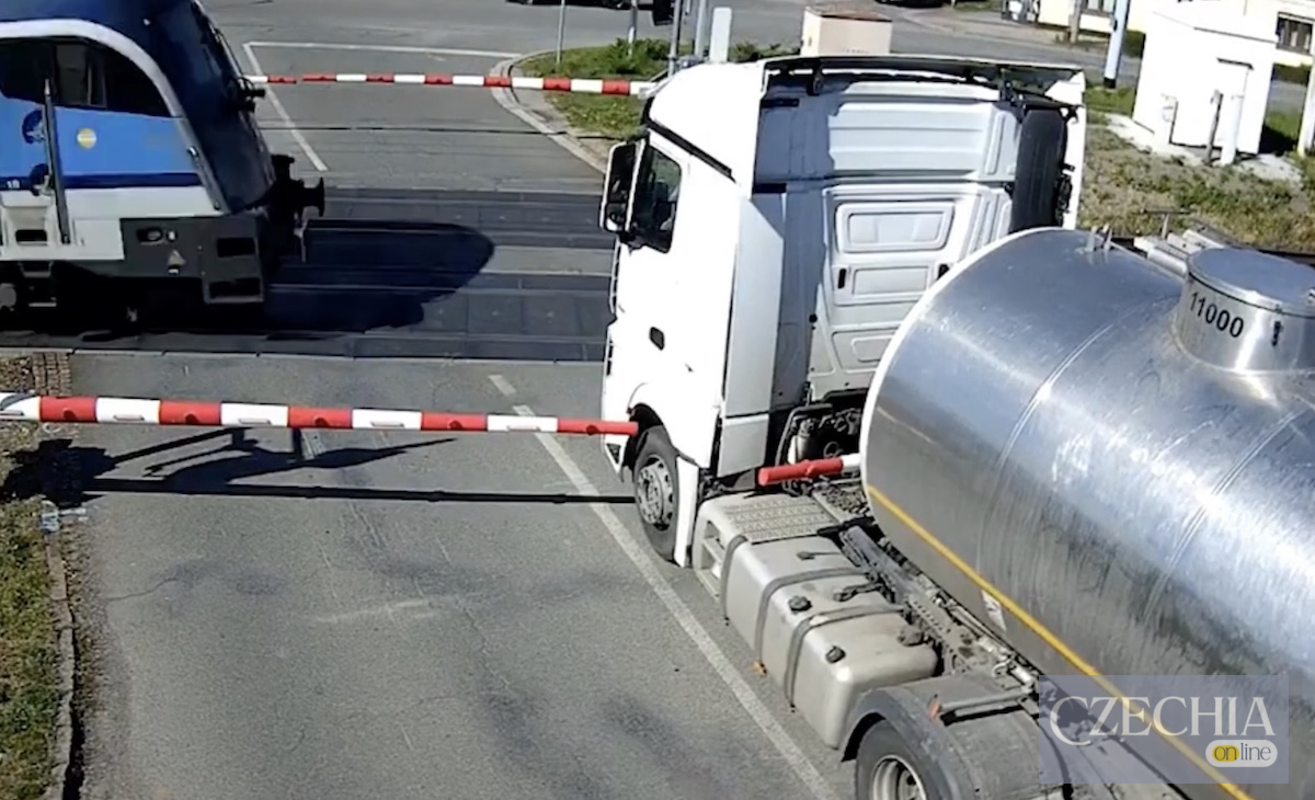 Біла фура з металевою цистерною стоїть затиснута шлагбаумом на залізничному переїзді впритул до потяга, що проїжджає повз.