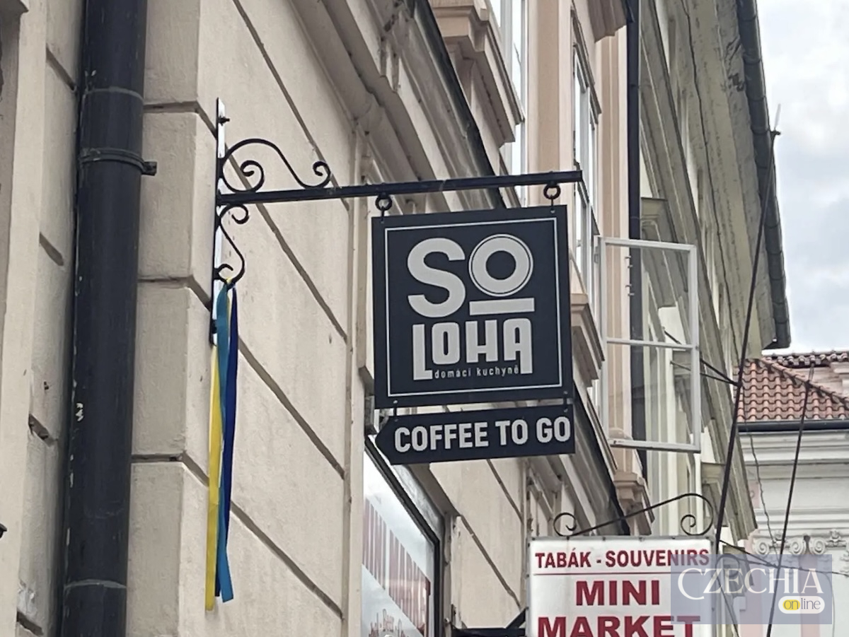 Чорна вивіска українського кафе Soloha на фасаді будівлі у центрі Праги. До кріплення прив'язана синьо-жовта стрічка. Матеріал про ціни в закладі.