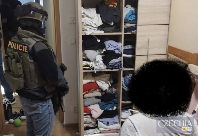 Озброєний поліцейський спецназу в тактичному спорядженні стоїть у кімнаті поруч із шафою під час обшуку. На передньому плані сидить затримана особа, обличчя якої приховане.