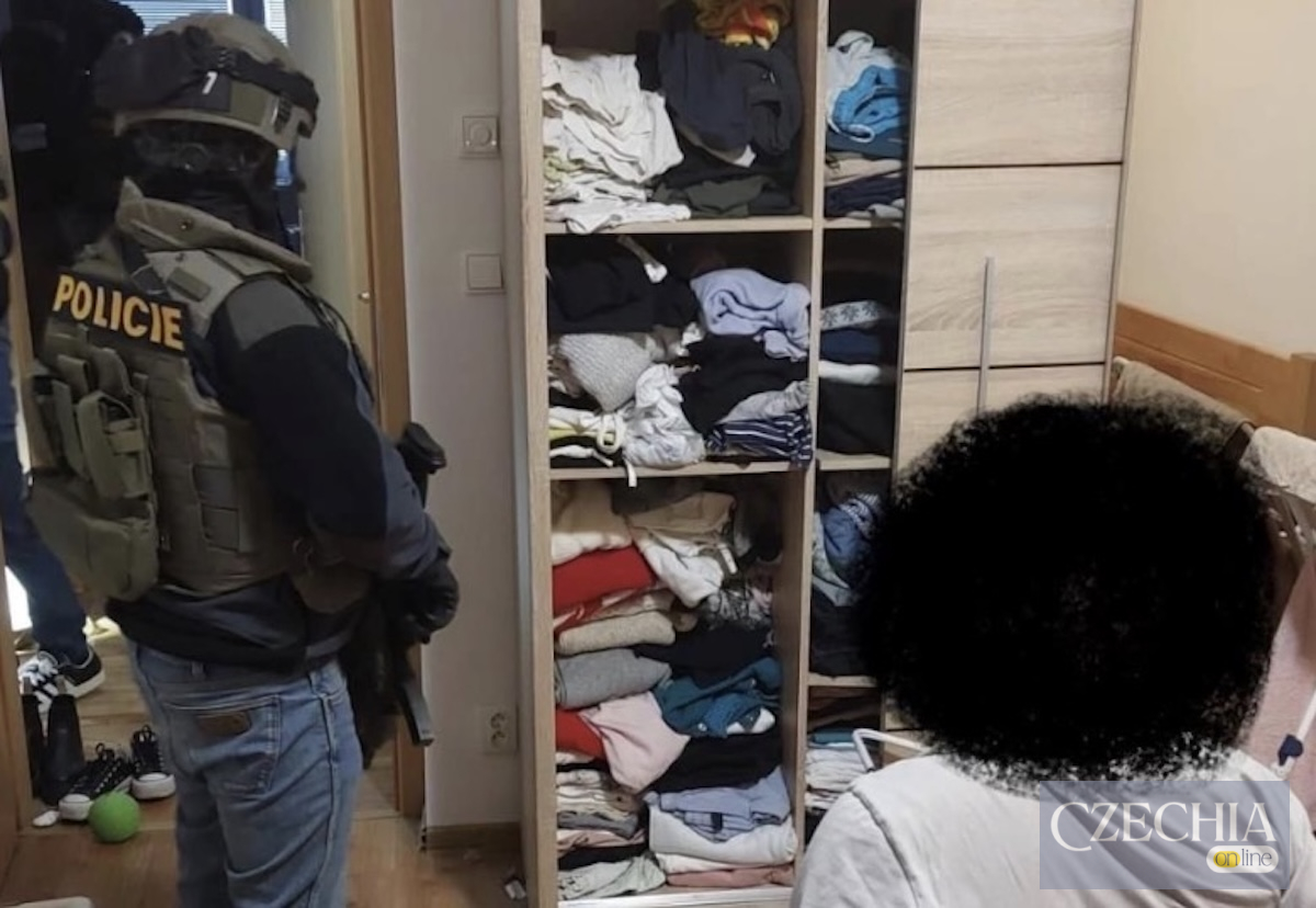 Озброєний поліцейський спецназу в тактичному спорядженні стоїть у кімнаті поруч із шафою під час обшуку. На передньому плані сидить затримана особа, обличчя якої приховане.